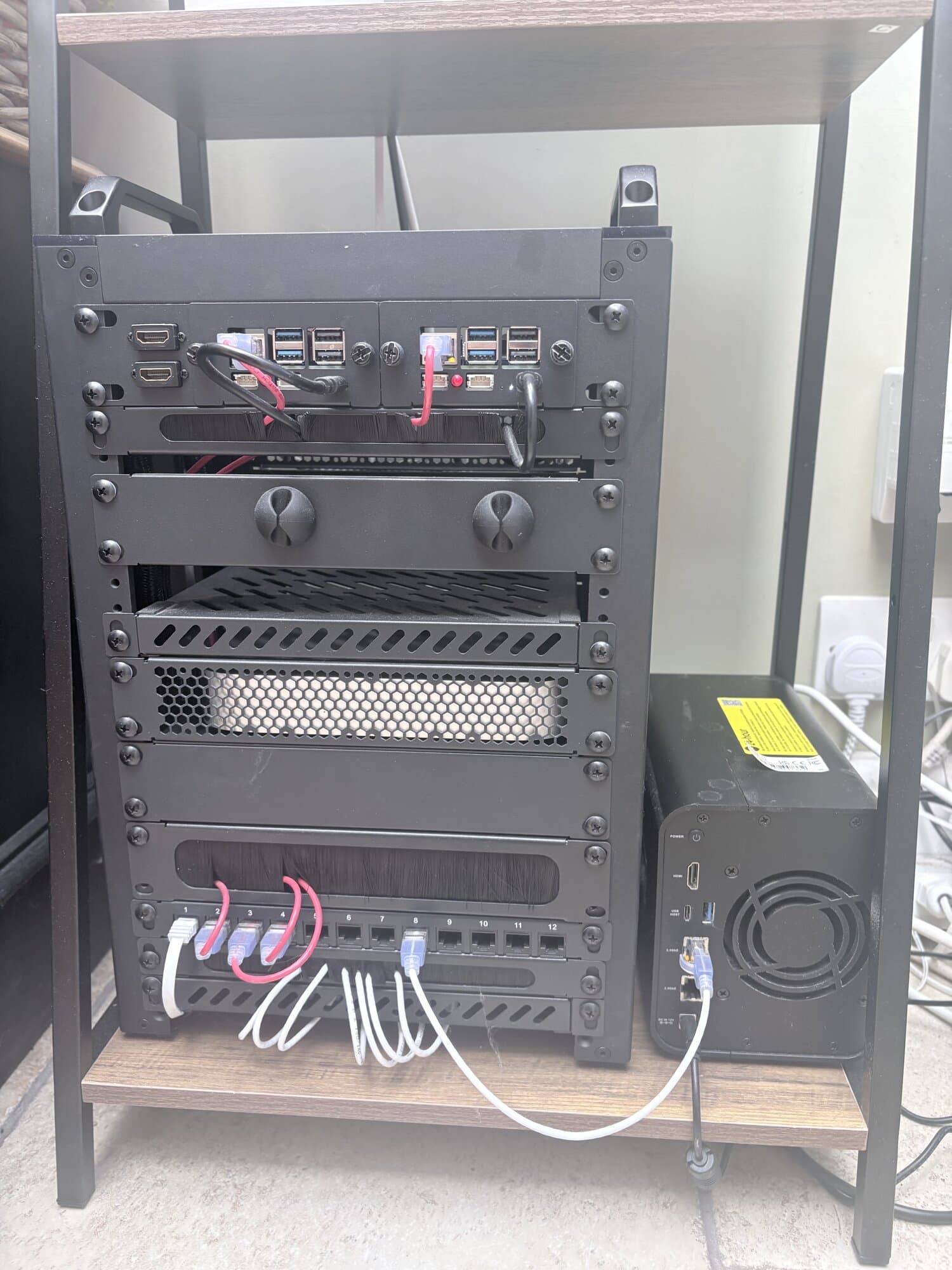 A small black equipment rack on a wooden shelf. Two Raspberry Pis are mounted in the top panel, a patch panel with a handful of network cables sits at the bottom, and a NAS stands next to the rack.
